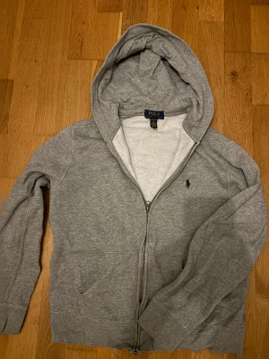 Grå Polo Ralph Lauren Zip Hoodie (14-16)  - Snygg hoodie från Polo Ralph Lauren i grå färg. Storleken är 14-16 (ungefär 160-170). Hoodien är i mycket bra kvalite och perfekt nu till den kalla vintern. Skriv om ni har frågor!