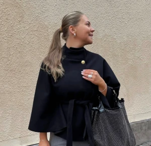 Svart kappa med bälte och guldknapp - Svart kappa med hög krage, breda ärmar och snyggt bälte i midjan. Guldknapp vid halsen ger en cool detalj. Insidan har ljusrosa foder som syns vid ärmsluten. Perfekt för dig som gillar stilrena och trendiga plagg. Poncho 