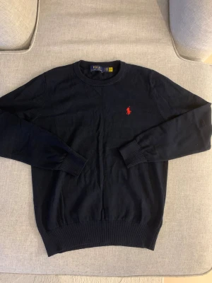 Mörkblå tröja från Polo Ralph Lauren - Snygg mörkblå tröja från Polo Ralph Lauren med klassisk röd logobrodering på bröstet. Tröjan har rund hals, långa ärmar och ribbade muddar vid ärmslut och nederkant. Tillverkad i mjuk bomull, perfekt för en chill och stilren look.