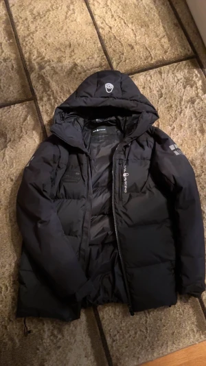 Sail Racing jacka  - Svart dunjacka från Sail Racing med huva och snygga logodetaljer på bröstet och ärmen. Jackan är fodrad med Allied Feather + Down 500 och har dragkedja framtill. Perfekt för kalla dagar och har en stilren, sportig look.