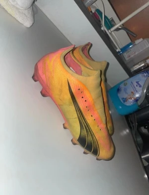 Gula och rosa fotbollsskor med svart detalj - Säljer ett par fotbollsskor från Puma i en riktigt fet färgkombo med gul och rosa bas samt svarta grafiska detaljer på sidan. Skorna har platt sula med dobbar och är tillverkade i syntetmaterial för grym komfort och lätt vikt. Perfekt för dig som vill sticka ut på planen.
