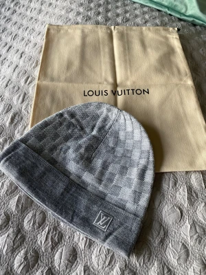 Grå Louis Vuitton mössa - Snygg grå mössa från Louis Vuitton med klassiskt rutigt mönster och LV-logga framtill. Mössan är stickad och har en bred uppvikt kant för extra stilpoäng. Perfekt accessoar för dig som gillar exklusiva märken och vill hålla dig varm med stil.