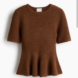 Brun stickad topp med volangkant - Superfin brun stickad topp med kort ärm och rund halsringning. Modellen har en figurnära passform och en snygg volangkant nedtill som ger en peplum-effekt. Perfekt att styla med både jeans och kjol för en trendig look.
