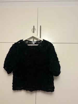 Svart blommig blus med kort ärm - Supersnygg svart blus från H&M med 3D-blommor över hela tyget. Blusen har rund halsringning och korta ärmar, perfekt för dig som gillar unika detaljer. Materialet är lätt och transparent med blommiga applikationer som ger en elegant look💕💕