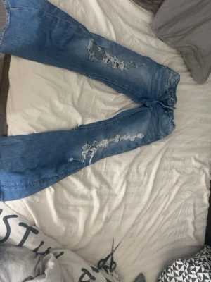 Blå ripped jeans från Gina Tricot - Säljer ett par ljusblå jeans från Gina Tricot med snygga slitningar och hål längs benen. Modellen har klassisk femficksdesign, knapp och dragkedja framtill samt justerbar midja. Perfekt för dig som gillar en trendig och avslappnad look.