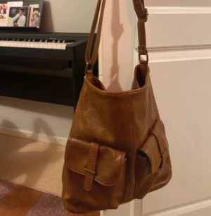 Brun axelväska med fickor i skinn - Säljer en brun axelväska i skinn med två stora fickor framtill och justerbar rem. Väskan har ett rymligt huvudfack med svart innerfoder och en mindre innerficka med dragkedja. Perfekt för dig som vill ha plats för allt du behöver under dagen.