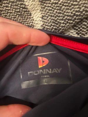 Blå Donnay padeltröja med retrotryck - Snygg mörkblå t-shirt från Donnay Padel i storlek L. Tröjan har ett stort vitt Donnay-tryck och en retroinspirerad rand i rött, orange och gult över bröstet. Tillverkad i ett lätt och syntetiskt material som passar perfekt för sport och träning.