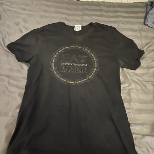 Svart EA7 Emporio Armani t-shirt - Svart t-shirt från EA7 Emporio Armani med rund hals och korta ärmar. Framsidan har ett stort, cirkulärt tryck med texten 'EA7 Emporio Armani Milano' i silverfärgade detaljer. Materialet känns mjukt och är troligtvis bomull, perfekt för en clean och stilren look.