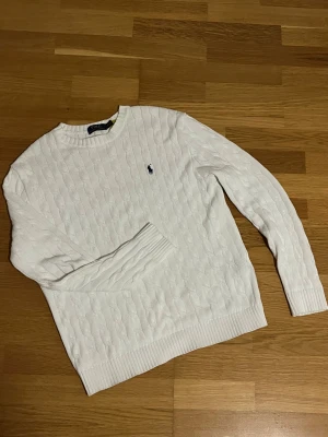 Polo Ralph Lauren Tröja - Vit kabelstickad tröja från Polo Ralph Lauren med klassisk rund hals och broderad marinblå logga på bröstet. Tröjan har ribbade muddar vid ärmslut och nederkant, långärmad och tillverkad i mjuk bomull. Perfekt för dig som gillar stilren och tidlös design.