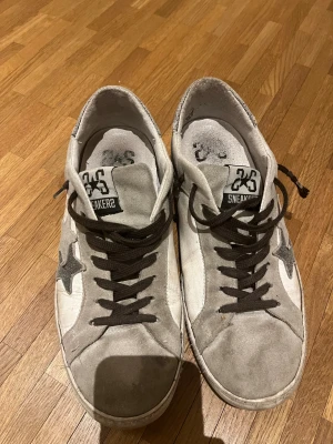 Gråvita sneakers från 2Star Sneakers - Säljer ett par gråvita sneakers från 2Star Sneakers med svart stjärndetalj på sidan. Skorna har svart snörning, rund tå och är tillverkade i mocka och skinn. Perfekta för dig som gillar en clean och stilren look med en twist.