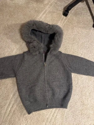 Alessa fur jacket  - Mysig grå stickad hoodie från S.Oliver med fluffig päls runt huvan. Tröjan har dragkedja framtill, ribbade muddar och långärmad design. Perfekt för kalla dagar när du vill vara både varm och snygg.