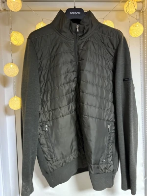 Mörkgrön quiltad pufferjacka Iceland XL - Snygg mörkgrön pufferjacka från Iceland i storlek XL. Jackan har quiltad front i syntetmaterial, stickade ärmar och två praktiska fickor med dragkedja. Hög krage och hel dragkedja framtill ger en cool look. Perfekt för dig som gillar stilren design.