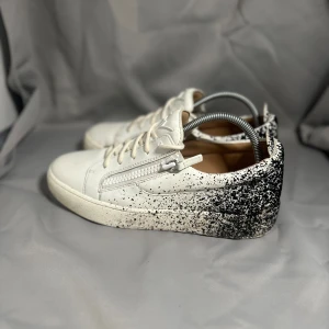 Vit-svarta sneakers från Giuseppe Zanotti - Säljer ett par snygga sneakers från Giuseppe Zanotti i vitt skinn med svart stänkmönster på hälen och sidorna. Skorna har snörning, dragkedja på sidan och rund tå. Perfekt för dig som gillar unik design och vill sticka ut med dina skor. Storlek 42,5 i bra skick 