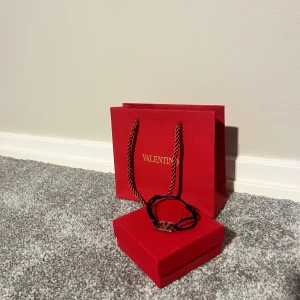  blått armband med Valentino-logga - Snyggt armband från Valentino med justerbart blått band och en silverfärgad V-logga i metall. Kommer i original röd box, tygpåse och presentpåse med gulddetaljer. Perfekt accessoar för att lyfta din outfit med en lyxig touch. Ena spänningen är lite lös. Nytt pris 2100kr