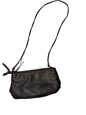 Svart axelväska i skinn från Åhléns - Snygg svart axelväska i äkta skinn från Åhléns med stilren design och praktisk dragkedja upptill. Väskan har en smidig axelrem och är perfekt för att bära dina viktigaste grejer. Enkel, klassisk och passar till många olika outfits.