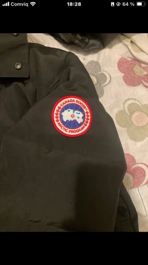 Svart Canada Goose dunjacka med päls - Svart dunjacka från Canada Goose med klassisk röd och vit logga på ärmen. Jackan har huva och medföljande beige pälsdetalj, samt knappar och dragkedja framtill. Perfekt för kalla vinterdagar och har en riktigt schysst streetstil.