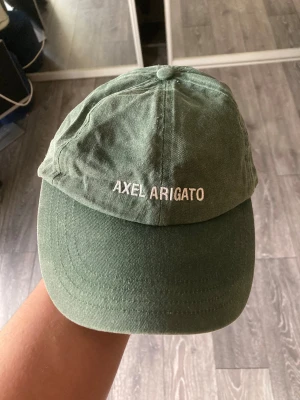 Grön keps från Axel Arigato - Snygg grön keps från Axel Arigato med broderad logga framtill. Kepsen är tillverkad i 100% bomull och har en klassisk böjd skärm. Perfekt accessoar för att lyfta din outfit med en clean och stilren look.