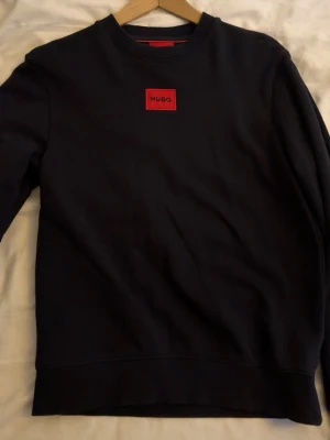 Mörkblå sweatshirt från Hugo Boss XS - Mörkblå sweatshirt från Hugo Boss i storlek XS. Tröjan har ett rött Hugo-märke på bröstet och klassisk rund hals. Materialet är mjukt och känns som bomull, med ribbade muddar vid ärmslut och nederkant. Perfekt för en clean och stilren look.
