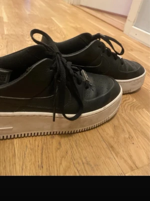 Svarta Nike Air Force 1 sneakers - Säljer ett par svarta Nike Air Force 1 sneakers med klassisk vit platt sula och snörning. Skorna har rund tå och är tillverkade i skinn med perforerade detaljer på tån. Snygg och tidlös modell som passar till streetwear och casual outfits.