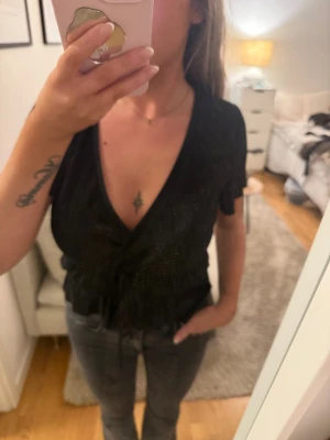 Svart omlottopp med snörning - Snygg svart omlottopp med korta ärmar och djup v-ringning.  tyg och knytband i midjan som ger en cool croppad look. Perfekt för dig som gillar att sticka ut med stilrena detaljer och lite extra shine.