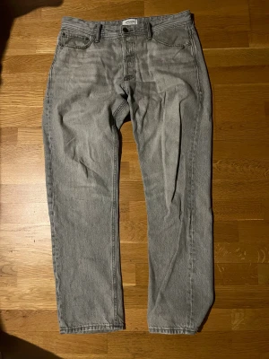 Grå raka jeans från Jack & jones - Snygga grå jeans från Jack & jones i klassisk rak modell. Jeansen har fem fickor, knapp och dragkedja framtill samt bälteshällor. Materialet är slitstarkt denim och färgen är ljusgrå med en cool tvättad look.