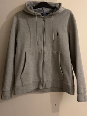 Polo ralph lauren Grå zip hoodie - Grå zip-hoodie i nyskick, knappt använd. Superskön insida i borstad fleece som är varm och mjuk mot huden. Storlek L på etiketten men passar även M bra (normal/rymlig passform). Inga fläckar, noppor eller slitage. Snygg, tidlös och perfekt nu till hösten!