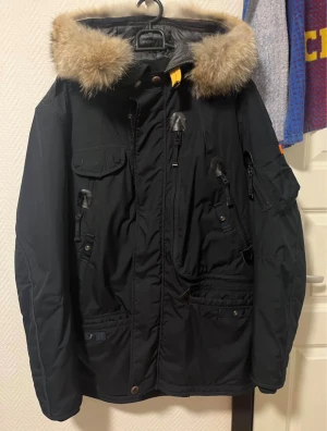 Svart Parajumpers vinterjacka med päls - Grym svart Parajumpers Masterlight vinterjacka med äkta päls på huvan, flera fickor och snygga detaljer som patch på ärmen och gul rem baktill. Jackan har dragkedja, knappar och är perfekt för kalla dagar. Slim fit-modell med lång ärm och robust känsla.