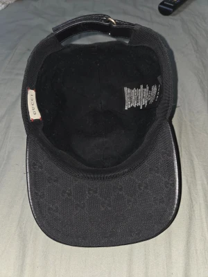 Svart Gucci keps med GG-mönster - Säljer en svart keps från Gucci med klassiskt GG-monogram över hela tyget. Kepsen har en böjd skärm och justerbart spänne bak i svart läder. På sidan finns en grön och röd rand som ger extra Gucci-vibe. Materialet är canvas och detaljer i läder. Chippet finns kvar såklart är den äkta. 