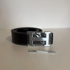 Svart skärp med H-spänne - Snyggt svart skärp från H med ett stort H-format spänne i silverfärgad metall. Skärpet är gjort av präglat skinn med vita sömmar längs kanterna för en stilren look. Perfekt accessoar för att lyfta din outfit.