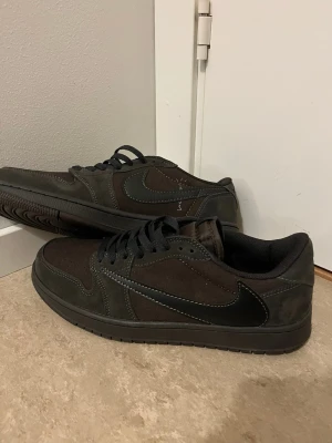 Nike Jordan 1 Low Travis Scott - Säljer ett par Nike Jordan 1 Low Travis Scott i mörkbrunt mocka med svarta detaljer och omvänd swoosh. Skorna har svarta snören, diskret broderad text och klassisk Jordan-sula. Perfekta för dig som gillar streetwear och unika sneakers.