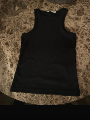 Svart ribbad linnetopp - Säljer en svart ribbad linnetopp med ärmlös design och rund hals. Toppen har en slim passform och är perfekt för att styla med jeans eller kjol. Materialet känns mjukt och stretchigt, vilket gör den skön att bära hela dagen.