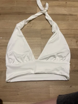 Vit halterneck topp med knyt - Säljer en vit halterneck topp med bred resår under bysten och knyt i nacken. Toppen är ärmlös och har en djup v-ringning. Tillverkad i mjuk och stretchig polyesterblandning som sitter snyggt på kroppen.