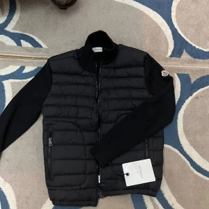 Moncler Cardigan jacka ny - Storlek S Aldrig använd Svart pufferjacka från Moncler med quiltad front och stickade ärmar. Jackan har hög krage, dragkedja framtill och två sidofickor med zip. Moncler-logga på vänster ärm. Perfekt för dig som vill ha en clean och stilren look.