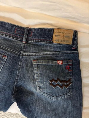 Lowaist jeans  - Säljer ett par skitsnygga blå jeans från Lowage med coola röda sömmar och detaljer. Låg midja och klassisk femficksmodell. Snyggt mönstrad tvätt på framsidan och broderad bakficka med zickzack. Perfekt för dig som gillar streetstyle.