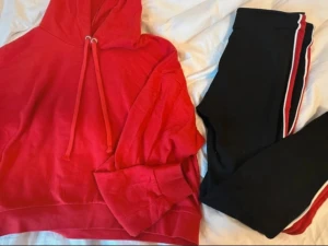 SET! Leggings med hoodie från H&M - Snygg röd croppad hoodie med huva och snörning framtill. Svarta leggings med en röd och vit rand längs utsidan av benen. De har en tight passform och hög midja, vilket ger en sportig och chill look. TRÖJAN ÄR I STORLEK M OCH BYXAN I STORLEK S