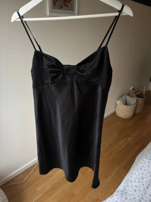Svart satin festklänning nyår jul - Så fin miniklänning perfekt för jul/nyår! Som en rosett vid bröstet. Köpt second hand, står inget märke. Knappt använd , jättebra skick. Banden går att justera. Passar till allt, så användbar 🪩 