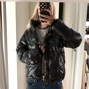 Svart pufferjacka från Bershka - Säljer en svart, glansig pufferjacka från Bershka i storlek S. Jackan har hög krage, dragkedja framtill och två sidofickor. Den är oversized med breda ärmar och tillverkad i syntetmaterial som ger en cool, modern look.