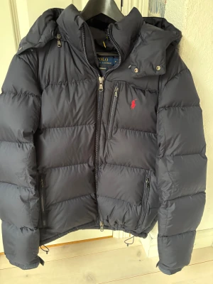 Mörkblå dunjacka från Polo Ralph Lauren - Säljer en mörkblå dunjacka från Polo Ralph Lauren med röd logga på bröstet. Jackan har huva, flera dragkedjefickor och justerbar nederkant med snörning. Perfekt för kalla dagar och har en schysst, puffig look som är riktigt trendig. Skick (10/10) Storlek (M) 