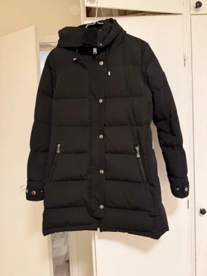 Svart lång dunjacka Hollies - Säljer en svart lång dunjacka från Hollies i storlek 38. Jackan har huva med snörning, dragkedja och knappar framtill samt två fickor med dragkedja. Snygg quiltad design och detaljer vid ärmslut. Perfekt för kalla dagar, har även kemtvätta den så den är helt ren