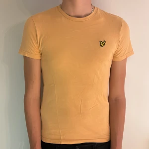 Orange t-shirt från Lyle & Scott - Snygg orange t-shirt från Lyle & Scott med klassisk rund hals och korta ärmar. Bröstet pryds av den ikoniska broderade örnloggan i gult och svart. Skön passform och tillverkad i mjuk bomull, perfekt för en clean och stilren look.