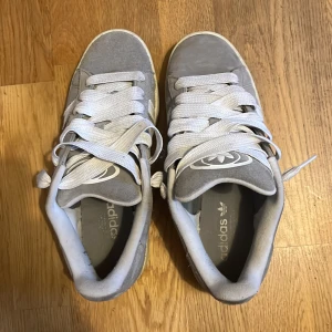 Grå Adidas Campus sneakers i mocka - Säljer ett par grå Adidas Campus sneakers med klassiska vita ränder på sidorna och vit sula. Skorna har breda vita skosnören och ovandel i mjuk mocka. Perfekta för dig som gillar en clean och tidlös stil. Gummisula med bra grepp.