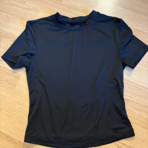 Svart sportig t-shirt i syntet - En svart, enkel och sportig t-shirt i syntetmaterial. Modellen är kortärmad med rund halsringning och har en slät, lätt glansig yta som passar perfekt till träning eller aktiva dagar.