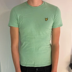 Mintgrön t-shirt från Lyle & Scott - Fräsch mintgrön t-shirt från Lyle & Scott med klassisk rund hals och korta ärmar. På bröstet sitter den ikoniska gula fågeln som broderad detalj. T-shirten är gjord i mjuk bomull och sitter skönt och snyggt mot kroppen.