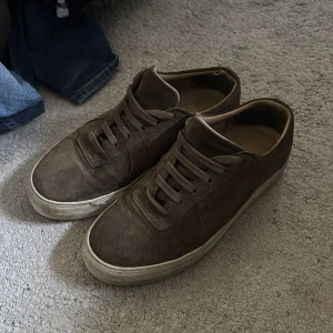 Bruna sneakers från Axel Arigato mocka - Snygga bruna sneakers från Axel Arigato i mocka med klassisk låg profil och rund tå. Skorna har snörning och vit platt sula som ger en clean look. Perfekta för dig som gillar stilrena och minimalistiska sneakers med premiumkänsla.