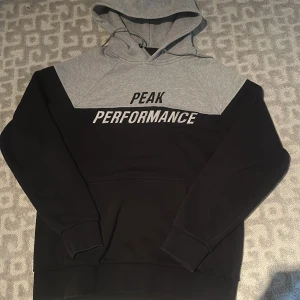 Grå och svart hoodie Peak Performance - Snygg hoodie från Peak Performance i storlek L. Tröjan är tvåfärgad med grått upptill och svart nedtill, har stor huva med snörning och en stor magficka. Broderad logga framtill. Perfekt för chill eller streetwear.