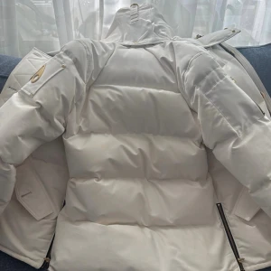 Vit dunjacka från Moose Knuckles - Snygg vit dunjacka från Moose Knuckles med guldfärgade detaljer, guldig logga på ärmen och lyxigt guldigt foder. Jackan har pälskantad huva, dragkedjor på ärmar och fram, samt flera fickor. Perfekt för kalla vinterdagar och riktigt statement-plagg. Pris går att diskutera 