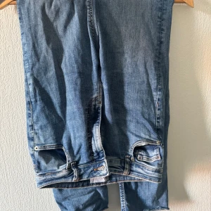 Blå jeans med slits från Zara - Säljer ett par blå jeans från Zara i klassisk denim med råa, avklippta benslut och slits vid ankeln. Jeansen har normal passform och snygga detaljer vid fickorna. Perfekta för en chill och trendig look.