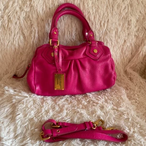 Rosa skinnväska från Marc Jacobs - Säljer en snygg handväska från Marc Jacobs i starkt rosa skinn med guldiga metalldetaljer. Väskan har dubbla handtag, avtagbar axelrem och coolt plisserad framsida. Perfekt för dig som vill sticka ut med en färgglad accessoar.
