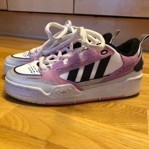 Adidas skor adi2000 - Snygga Adidas sneakers med klassiska svarta ränder, vit bas och detaljer i ljusrosa mocka. Skorna har vit snörning, svart innersida och coola rosa detaljer på sulan. Skorna är i använt men mycket bra skick då de endast använts några gånger men insidan är lite nopprig. Nypris är ca 1200 kr.