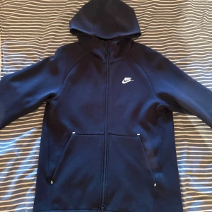 Mörk blå  Nike hoodie med dragkedja - Säljer en mörk blå  hoodie från Nike i storlek S. Tröjan har huva, hel dragkedja och två stora fickor framtill. Nike-loggan i vitt på bröstet ger en clean look. Perfekt för chill dagar eller när du vill ha en sportig stil.